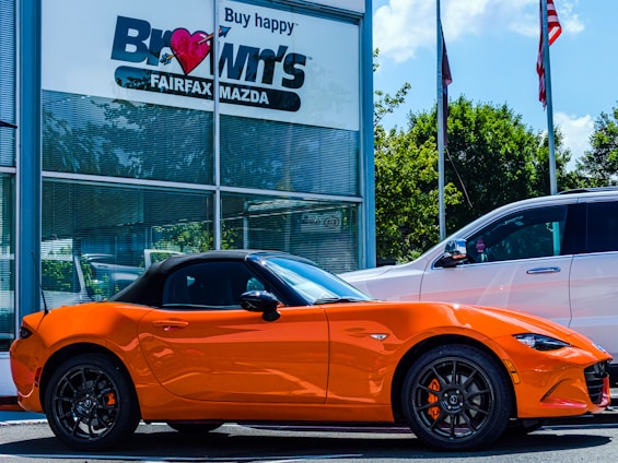 A bright orange convertible sports car is parked outside a car dealership. The building has a sign with 'Brown's FAIRFAX MAZDA' on it, and there are two flags flying nearby. A larger white SUV is also parked next to the sports car. Trees and a clear blue sky provide a backdrop.