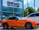 A bright orange convertible sports car is parked outside a car dealership. The building has a sign with 'Brown's FAIRFAX MAZDA' on it, and there are two flags flying nearby. A larger white SUV is also parked next to the sports car. Trees and a clear blue sky provide a backdrop.