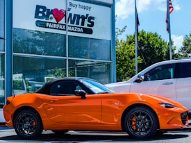A bright orange convertible sports car is parked outside a car dealership. The building has a sign with 'Brown's FAIRFAX MAZDA' on it, and there are two flags flying nearby. A larger white SUV is also parked next to the sports car. Trees and a clear blue sky provide a backdrop.