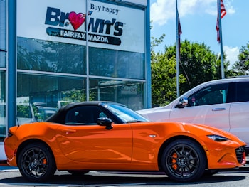 A bright orange convertible sports car is parked outside a car dealership. The building has a sign with 'Brown's FAIRFAX MAZDA' on it, and there are two flags flying nearby. A larger white SUV is also parked next to the sports car. Trees and a clear blue sky provide a backdrop.