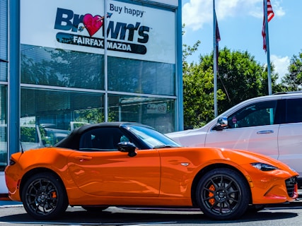 A bright orange convertible sports car is parked outside a car dealership. The building has a sign with 'Brown's FAIRFAX MAZDA' on it, and there are two flags flying nearby. A larger white SUV is also parked next to the sports car. Trees and a clear blue sky provide a backdrop.