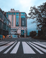 A multi-story commercial building stands at the end of an empty street with a crosswalk in the foreground. The building has large advertisements and various signs. There are trees and streetlights along the road, and a few people can be seen walking along the sidewalk. The sky appears cloudy and overcast.