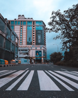 A multi-story commercial building stands at the end of an empty street with a crosswalk in the foreground. The building has large advertisements and various signs. There are trees and streetlights along the road, and a few people can be seen walking along the sidewalk. The sky appears cloudy and overcast.