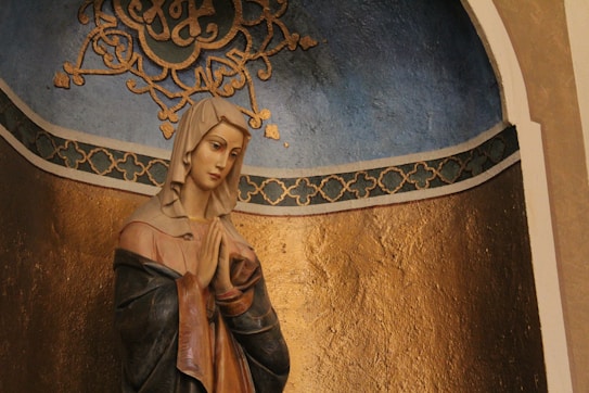 A statue of a religious figure, possibly the Virgin Mary, is positioned against a richly textured gold background. The statue features a serene expression, with hands clasped in prayer. It is adorned in a cloak with subtle shades of blue and pink. Above the figure, there is ornate detailing with a scripted design in gold against a muted blue backdrop.