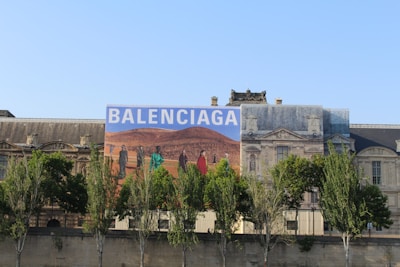 A large building with classic architecture, featuring an oversized advertisement with the word 'Balenciaga'. The advertisement depicts a desert landscape with people dressed in fashion-forward clothing.