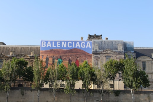 A large building with classic architecture, featuring an oversized advertisement with the word 'Balenciaga'. The advertisement depicts a desert landscape with people dressed in fashion-forward clothing.