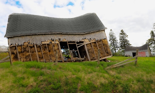 A dilapidated and leaning barn with visible hay inside, situated on a grassy field. The barn appears to have structural damage with missing and broken wood panels. In the background are several tall trees and another smaller, worn-out building.