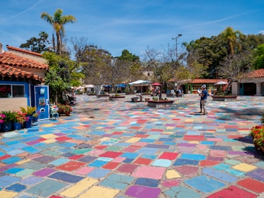 A vibrant outdoor plaza features a multicolored tiled ground, surrounded by trees and a few small buildings with red-tiled roofs. There are people casually walking and others seated under umbrellas in the distance. Potted plants and flowers add bursts of color, and palm trees are visible against a clear blue sky.