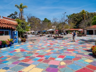 A vibrant outdoor plaza features a multicolored tiled ground, surrounded by trees and a few small buildings with red-tiled roofs. There are people casually walking and others seated under umbrellas in the distance. Potted plants and flowers add bursts of color, and palm trees are visible against a clear blue sky.