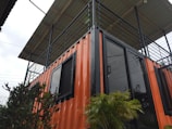 A structure made from a repurposed shipping container with an orange exterior and black trim. The container is fitted with black-framed glass doors and windows. On top, there is a patio surrounded by a black metal railing and covered with a roof. There are green plants in front of the container, adding a touch of nature.