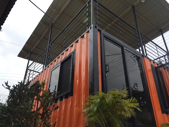 A structure made from a repurposed shipping container with an orange exterior and black trim. The container is fitted with black-framed glass doors and windows. On top, there is a patio surrounded by a black metal railing and covered with a roof. There are green plants in front of the container, adding a touch of nature.