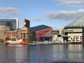 In a vibrant waterfront cityscape, a mix of modern and historic architecture lines the shore. A prominent hard rock cafe sign with a guitar shape stands out among the red-brick buildings. A bright red ship named Chesapeake is docked in the calm blue waters. The scene includes a geometric glass pyramid and a colorful building with square patterns, creating an eclectic urban atmosphere.