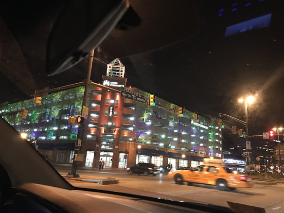 A nighttime urban setting features a brightly lit multi-story building with colorful lights illuminating its facade. The structure is identified as a medical facility by the signage. Yellow cabs and other cars are visible on the street, indicating typical city traffic.