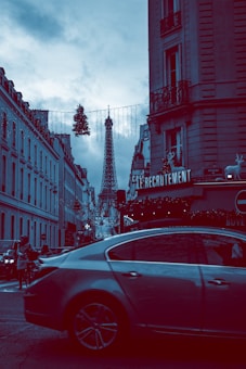 A street scene in Paris with the Eiffel Tower prominently visible in the background. The street is lined with classic Parisian buildings adorned with Christmas decorations, including lights and wreaths. A car is passing by in the foreground, and pedestrians are visible on the sidewalks. The sky is overcast, adding a moody atmosphere.