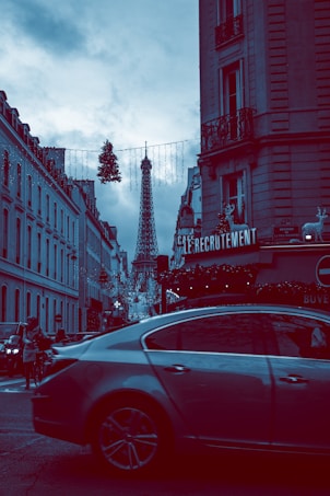 A street scene in Paris with the Eiffel Tower prominently visible in the background. The street is lined with classic Parisian buildings adorned with Christmas decorations, including lights and wreaths. A car is passing by in the foreground, and pedestrians are visible on the sidewalks. The sky is overcast, adding a moody atmosphere.