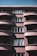 A multi-story residential building with a modern architectural style is depicted. The building features curved balconies with shadow patterns created by sunlight. The facade is primarily composed of large windows, some of which reflect the sky and clouds. The structure is painted in muted brown and beige tones, with a clear sky visible above.