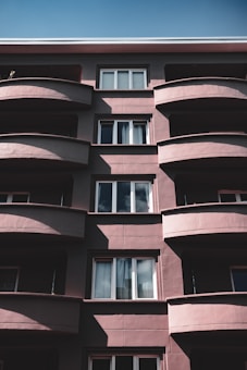 A multi-story residential building with a modern architectural style is depicted. The building features curved balconies with shadow patterns created by sunlight. The facade is primarily composed of large windows, some of which reflect the sky and clouds. The structure is painted in muted brown and beige tones, with a clear sky visible above.