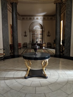 A sophisticated and elegant interior room with classical architecture. At the forefront, a round table with intricate golden details stands on a decorative tiled floor. In the background, two large marble columns frame the room, which is adorned with a bookshelf filled with volumes and flanked by chairs. Soft, ambient lighting casts gentle shadows, complementing the room's ornate details and luxurious furniture.