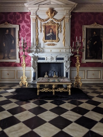 The image features an ornate and luxurious interior with a checkered marble floor and a grand fireplace adorned with intricate gold details. Above the fireplace is a portrait in a gold frame. The walls are covered with rich red wallpaper and flanked by two standing candelabras on either side. A decorative bench with a matching gold frame and embroidered cushion is placed in front of the fireplace.
