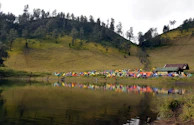 An aerial view of a large outdoor festival by a lake, showcasing vibrant tents and happy attendees.