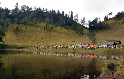 An outdoor scene features a large group of colorful tents pitched on a grassy area by a lake, with the reflection of the tents visible in the water. Surrounding the tents are lush green hills dotted with trees and a clear sky above.