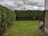 A small backyard featuring a neatly trimmed lawn bordered by dense green hedges and a wooden fence. The sky is overcast with grey clouds, and there is a bare tree visible above the fence. A brick house with a small stairway leading to a wooden porch is on the right.
