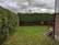 A small backyard featuring a neatly trimmed lawn bordered by dense green hedges and a wooden fence. The sky is overcast with grey clouds, and there is a bare tree visible above the fence. A brick house with a small stairway leading to a wooden porch is on the right.
