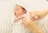 A newborn baby lies comfortably with a soft, white hat and clothing. Someone gently adjusts the baby's outfit with careful hands. The baby has a serene expression, with its mouth slightly open, indicating a moment of calm or rest.