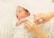 A newborn baby lies comfortably with a soft, white hat and clothing. Someone gently adjusts the baby's outfit with careful hands. The baby has a serene expression, with its mouth slightly open, indicating a moment of calm or rest.