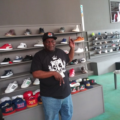 A man is standing in a shoe store, surrounded by displays of sneakers and caps. He is pointing upwards and smiling, wearing a black t-shirt with a graphic design and blue jeans. The shelves behind him hold a variety of colorful shoes, and there is a counter in front of him with several caps on display.