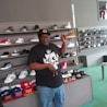 A man is standing in a shoe store, surrounded by displays of sneakers and caps. He is pointing upwards and smiling, wearing a black t-shirt with a graphic design and blue jeans. The shelves behind him hold a variety of colorful shoes, and there is a counter in front of him with several caps on display.
