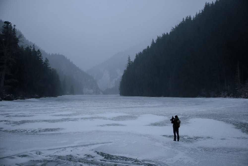 man standing on freezing body of water while facing trees and mountains