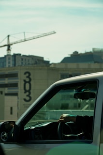 A person is driving a vehicle, with their hand resting on the steering wheel. The vehicle is in front of a building with a large number '303' displayed on its facade. A construction crane and modern buildings are visible in the background under a blue sky.