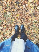 A person is standing among fallen autumn leaves, wearing black boots, black pants, and a denim jacket. The ground is covered with a thick layer of brown, orange, and yellow leaves.