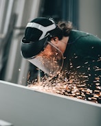A person wearing protective gear operates a welding or grinding tool, producing bright sparks. The person is focused on the task, with a clear face shield and ear protection.