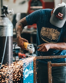 A person is using a yellow angle grinder to cut through metal, producing sparks flying in different directions. The individual is wearing a cap and a blue t-shirt with designs on it. This scene takes place in a workshop or industrial setting, with tools and equipment visible in the background.