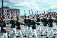 A group of uniformed individuals marching in formation along a street lined with historical buildings and ships in the background. They are wearing dark jackets, white trousers, and hats, carrying rifles on their shoulders. The setting suggests a ceremonial or formal event.