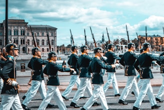 A group of uniformed individuals marching in formation along a street lined with historical buildings and ships in the background. They are wearing dark jackets, white trousers, and hats, carrying rifles on their shoulders. The setting suggests a ceremonial or formal event.