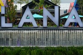 Large illuminated letters are situated in front of a stylish water fountain. In the background, colorful umbrellas and modern architecture are visible, adding a lively atmosphere to the scene.