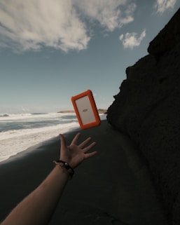 A hand is extended upward on a dark sandy beach, and an external hard drive with an orange protective case is in mid-air as if thrown or caught. The ocean waves are crashing in the background under a partly cloudy sky, with a rocky cliff on the right.