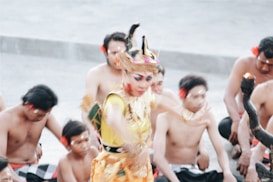 A traditional dancer dressed in colorful, ornate attire performs in front of a group of men wearing minimal clothing. The dancer's costume includes gold and yellow fabrics, and they are adorned with a decorative headpiece. The men sitting around are wearing checkered sarongs and have white facial paint, possibly for a cultural or ritualistic performance.