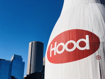 A large outdoor billboard featuring the word 'Hood' in white text on a red oval background stands prominently. The background consists of modern high-rise buildings with glass facades under a clear blue sky.