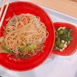 A vibrant red bowl filled with freshly prepared noodles, garnished with slices of tomato and greens, paired with a side dish of green leafy vegetables mixed with white beans, all placed on a white tray with wooden chopsticks.