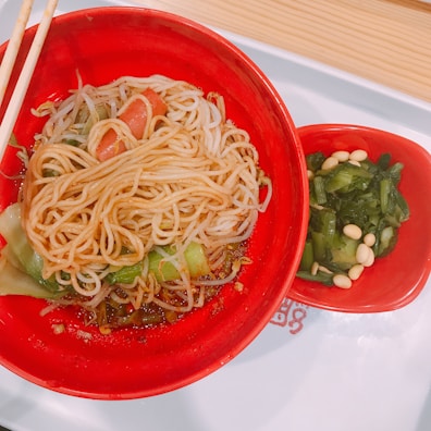A vibrant red bowl filled with freshly prepared noodles, garnished with slices of tomato and greens, paired with a side dish of green leafy vegetables mixed with white beans, all placed on a white tray with wooden chopsticks.