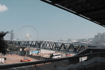 A cityscape featuring a large ferris wheel near the shoreline, with a bridge crossing over a busy road where taxis and a bus are visible. Skyscrapers are in the background, and a clear sky enhances the scene.