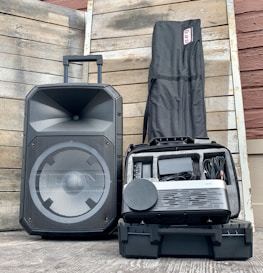 A large speaker with a handle is placed next to a protective case that contains electronic equipment, including cables and devices. The setup is arranged against a wooden wall background, and a black bag stands upright next to the case.