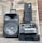 A large speaker with a handle is placed next to a protective case that contains electronic equipment, including cables and devices. The setup is arranged against a wooden wall background, and a black bag stands upright next to the case.