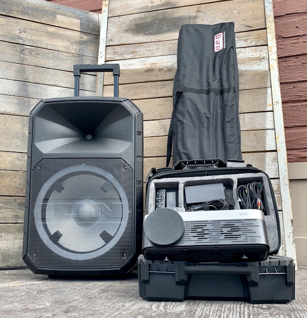 A large speaker with a handle is placed next to a protective case that contains electronic equipment, including cables and devices. The setup is arranged against a wooden wall background, and a black bag stands upright next to the case.