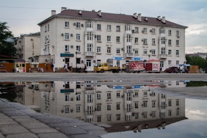 A large, multi-story residential building is reflected in a puddle on the pavement. The building features multiple windows and air conditioning units on its facade. In front of the building, there are several parked vehicles, including a yellow truck and a red van. People are walking along the sidewalk, and there are street vendors selling goods from carts.