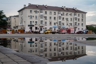 A large, multi-story residential building is reflected in a puddle on the pavement. The building features multiple windows and air conditioning units on its facade. In front of the building, there are several parked vehicles, including a yellow truck and a red van. People are walking along the sidewalk, and there are street vendors selling goods from carts.